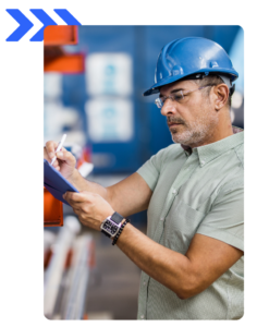 Industrial worker wearing a blue hard hat and safety glasses taking notes on a clipboard.