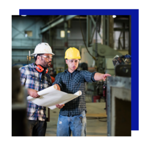 Two engineers in hard hats review blueprints in a fabrication facility.