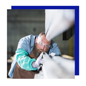 Bearded metalworker inspects a long beam for precision in a fabrication shop.