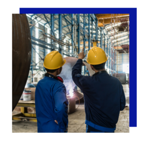 Two fabrication workers in safety gear coordinate inside a large metal workshop.